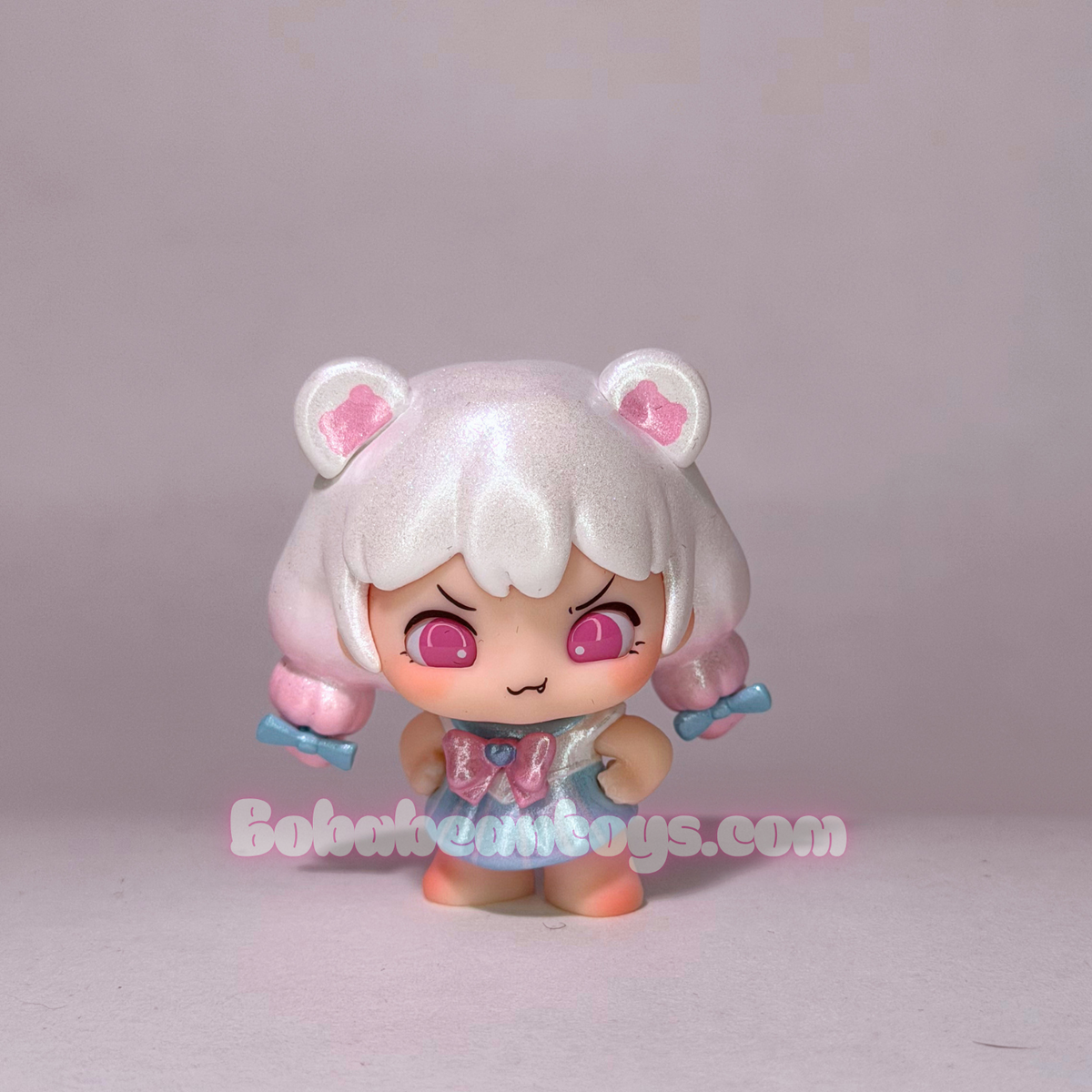 A small anime-style collectible figurine with short white hair, pink cat ears, and pink eyes, wearing a light blue and pink outfit. The figurine is photographed against a plain light gray studio background with a watermark reading 'Bobabeantoys.com