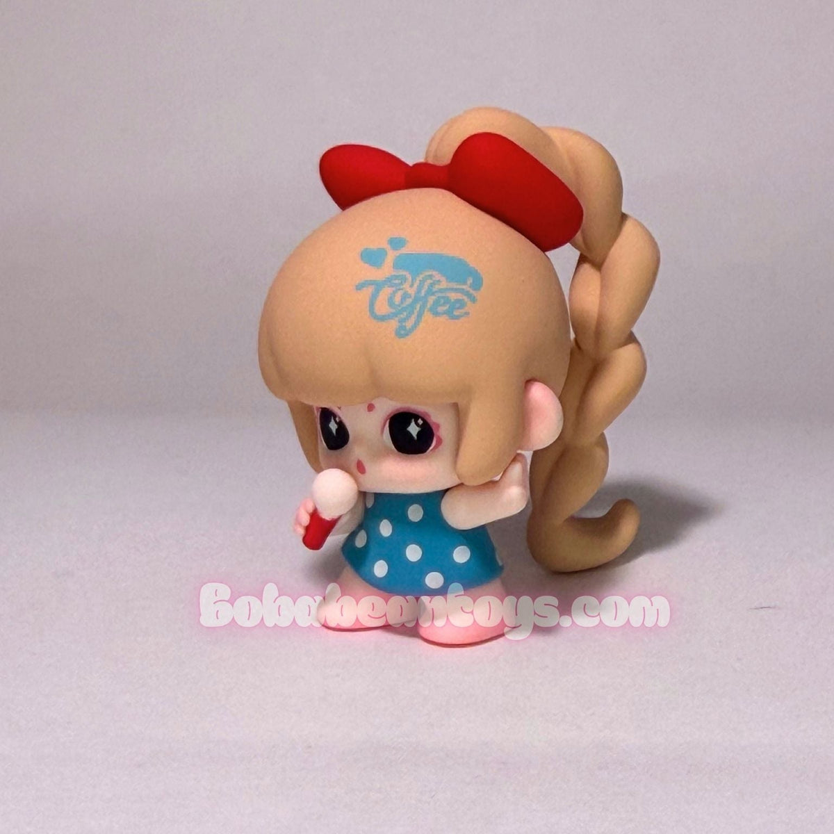 A small stylized doll figurine with a tan head, large black eyes, a red bow, braided hair, a blue polka-dot dress, and a red-and-white striped cup, photographed on a plain light gray background.
