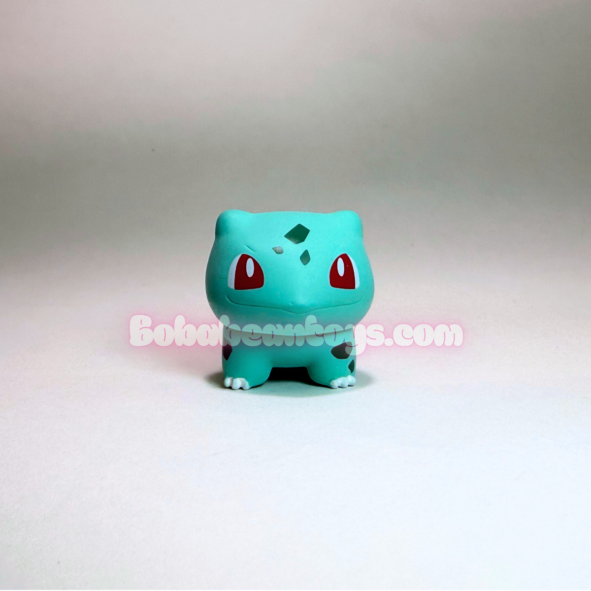 A small blue-green Bulbasaur figurine from Pokémon, standing against a plain light gray background. The figure has red eyes, a green badge on its forehead, and a pink watermark across the center reading 'Boboibeargus.