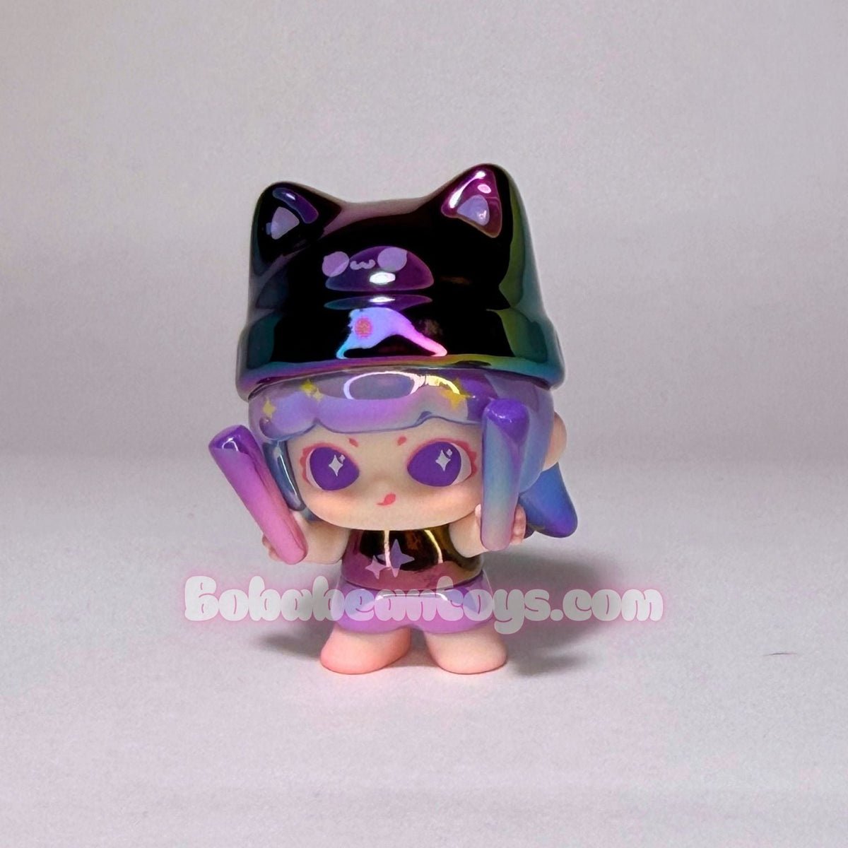 A small stylized toy figure with a glossy black cat-ear hat, purple goggles, and pastel pink and blue hair, standing against a plain light gray background. A faint 'BoholianToys.