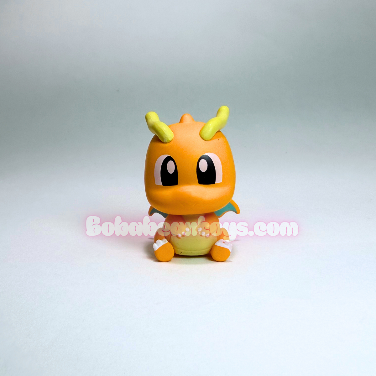 A small orange dragon figurine with yellow horns, large black eyes, and a yellow belly, centered on a plain light gray studio background. A faint 'BoboTos.
