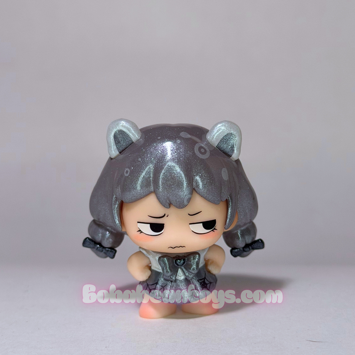 A small anime-style vinyl figure with short metallic silver-gray hair, cat-ear accessories, and a grumpy expression, standing against a plain light gray background. A faint bobabeantoys.com