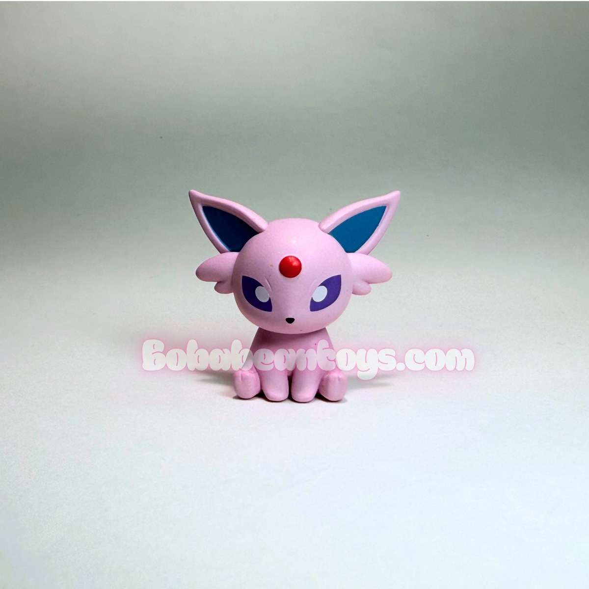 A small pink toy figurine resembling a cute cat-like Pokémon character with blue inner ears, purple eyes, and a red forehead gem, photographed against a plain light gray studio background. A semi-transparent 'Bobbeewo toys.