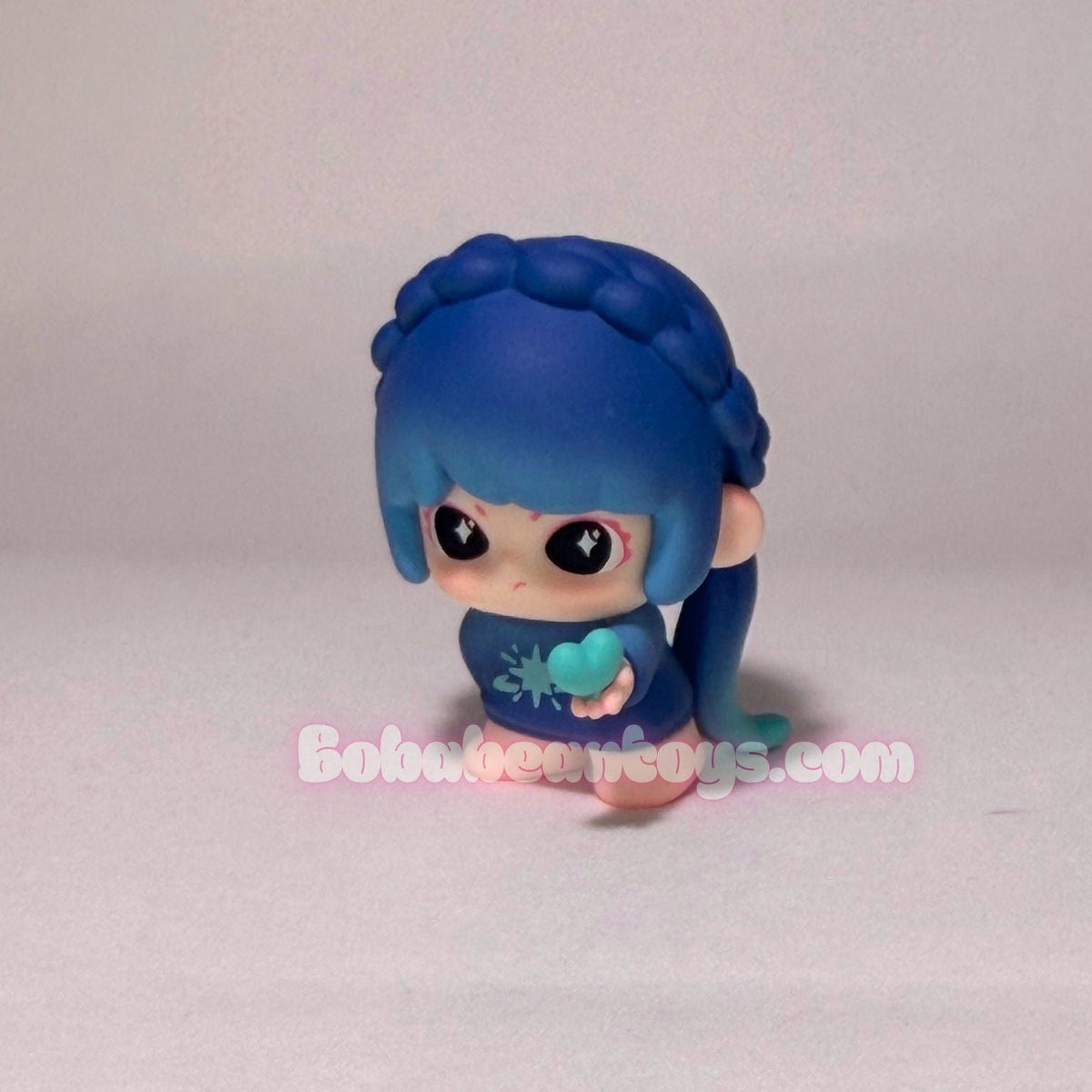 A small blue cartoon figurine with large eyes, braided hair, and a blue outfit holding a light blue heart, photographed against a plain light background. A watermark reading Boboobos Toys.