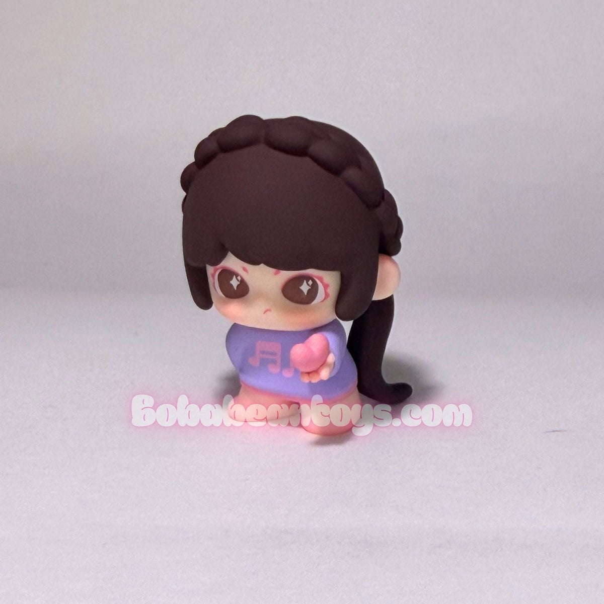 A small chibi-style toy figurine with dark brown hair, large brown eyes, and a purple outfit holding a pink heart, photographed against a plain light gray background. A watermark reading BobaBeanToys.