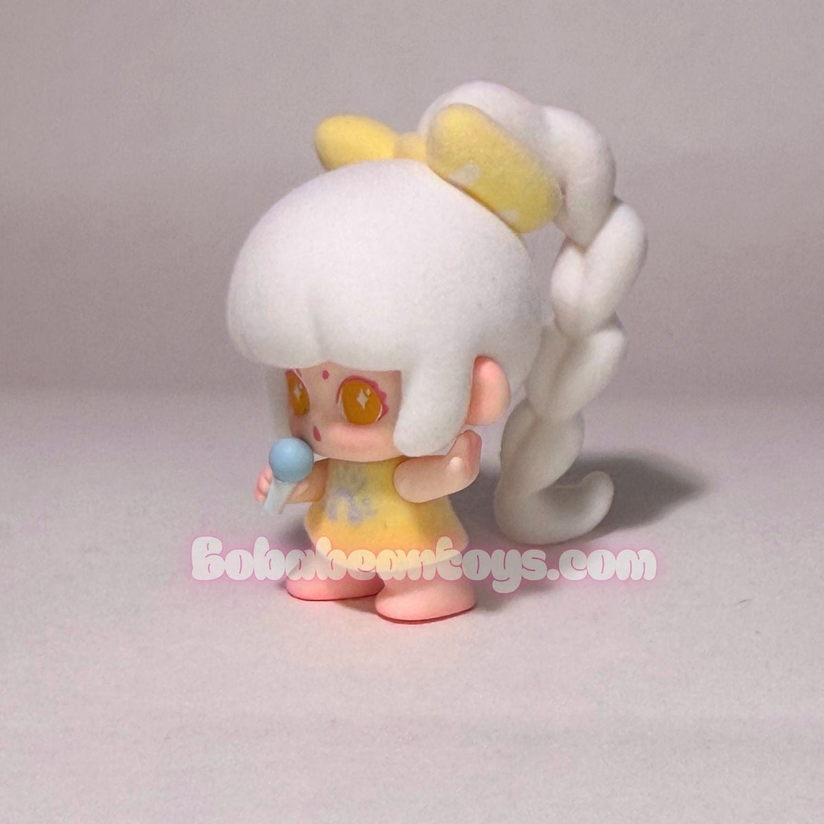 A small stylized doll figure with white hair in a high ponytail, yellow bow, yellow dress, and pink shoes, standing against a plain light gray background. A watermark reading BobabeansToys.