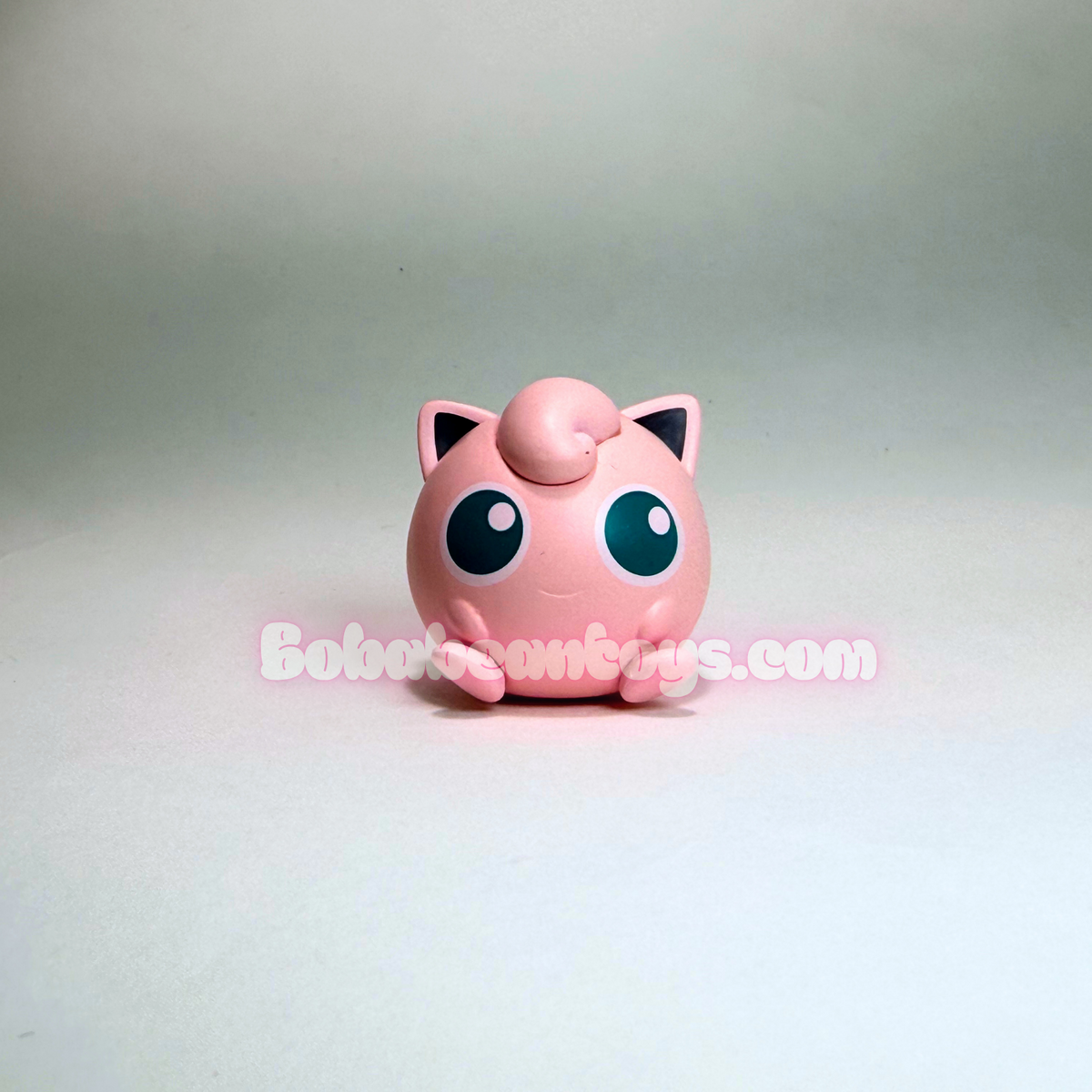 A small pink Jigglypuff toy figurine with large blue eyes, photographed against a plain light gray studio background. A semi-transparent 'Bob's Burgers.