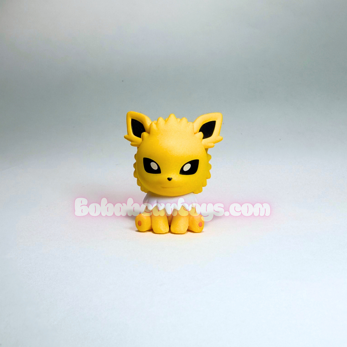 A small yellow Eevee toy figurine with black eyes and ears, standing on a white surface against a light gray background. A semi-transparent 'Bobatwixus.