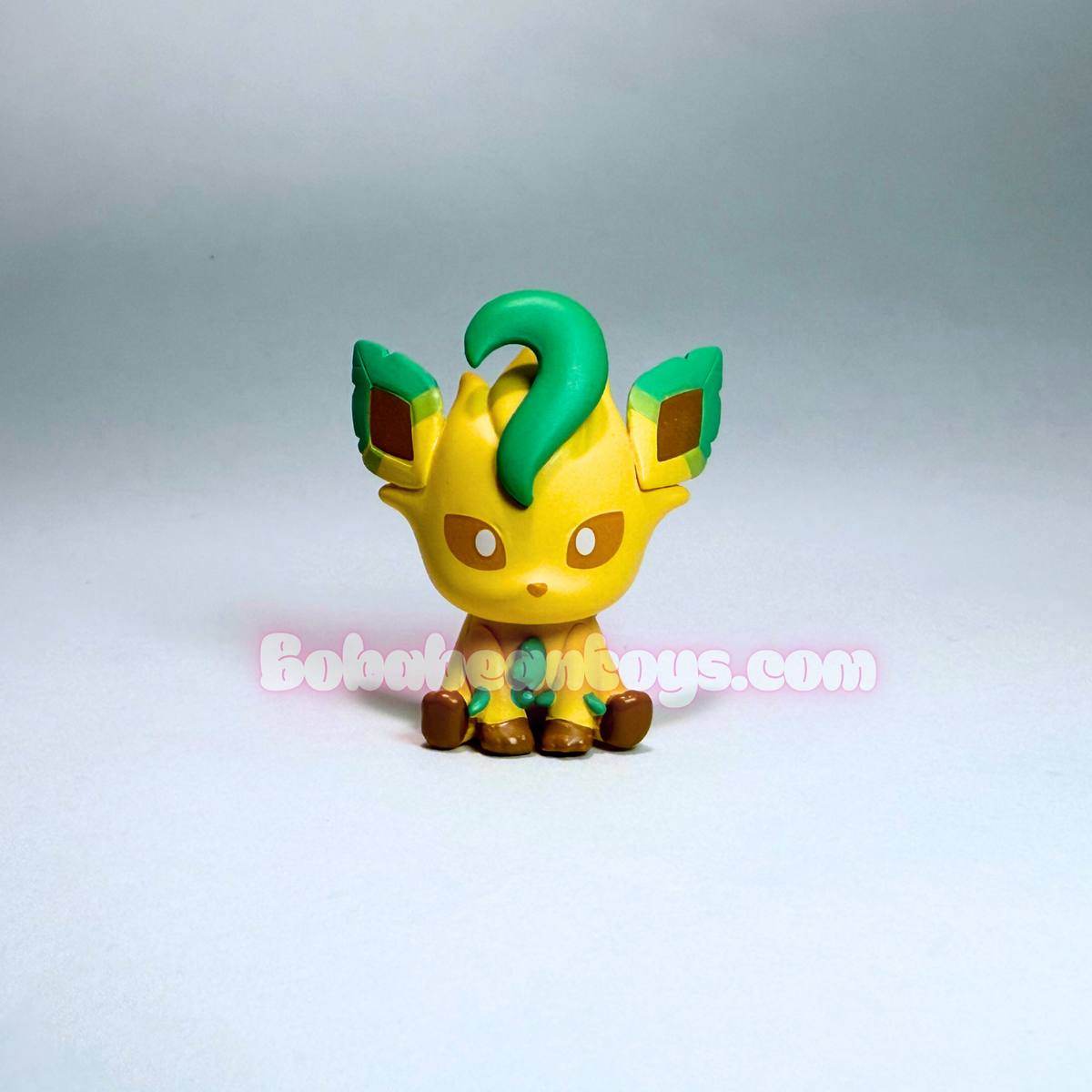 A small yellow and green toy figure resembling a cute Eevee character, photographed against a plain light gray studio background. A faint 'Bobaitoys.