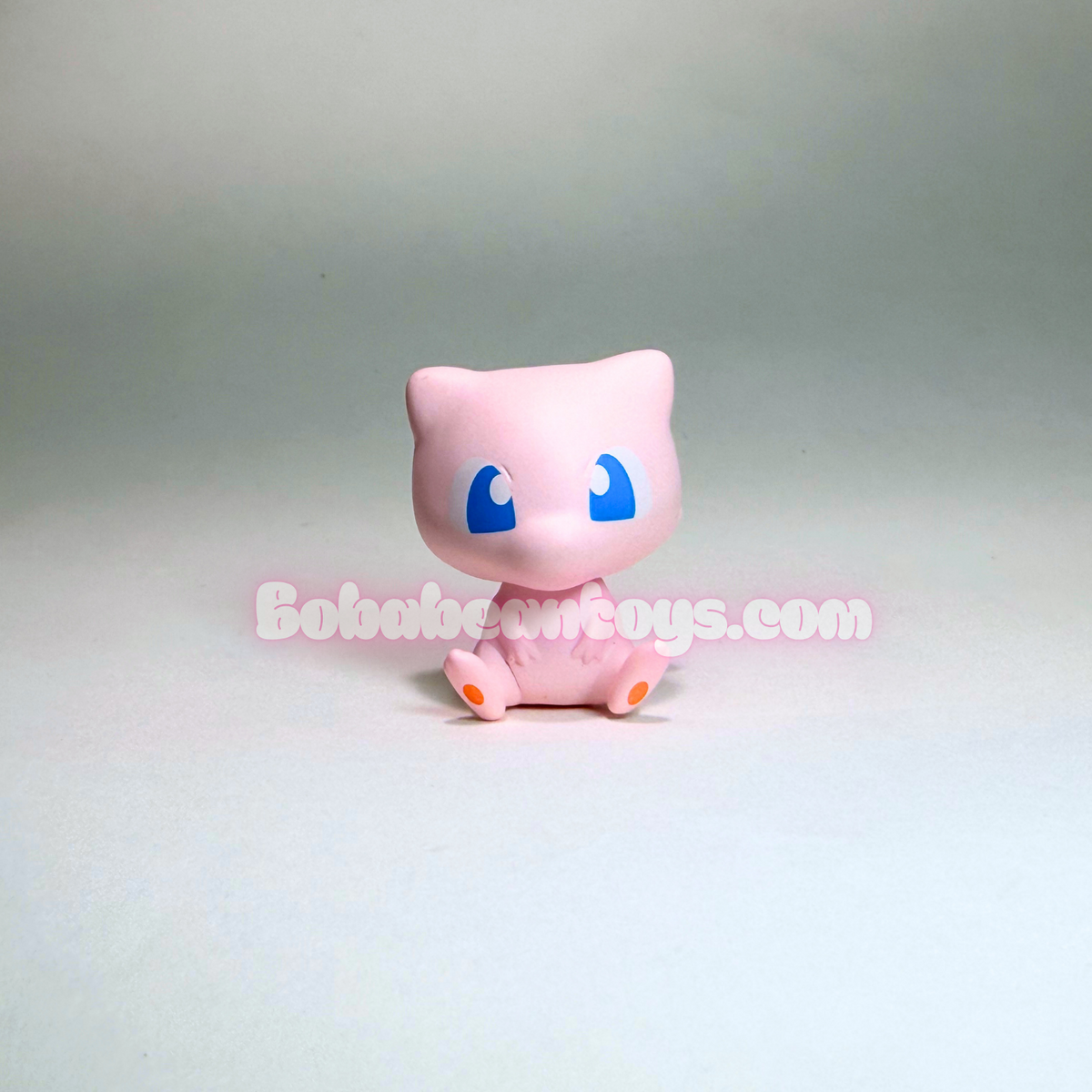 A small pink cat-like toy figure with large blue eyes and tiny orange feet, photographed against a plain light gray studio background. A semi-transparent 'BobbeanToys.