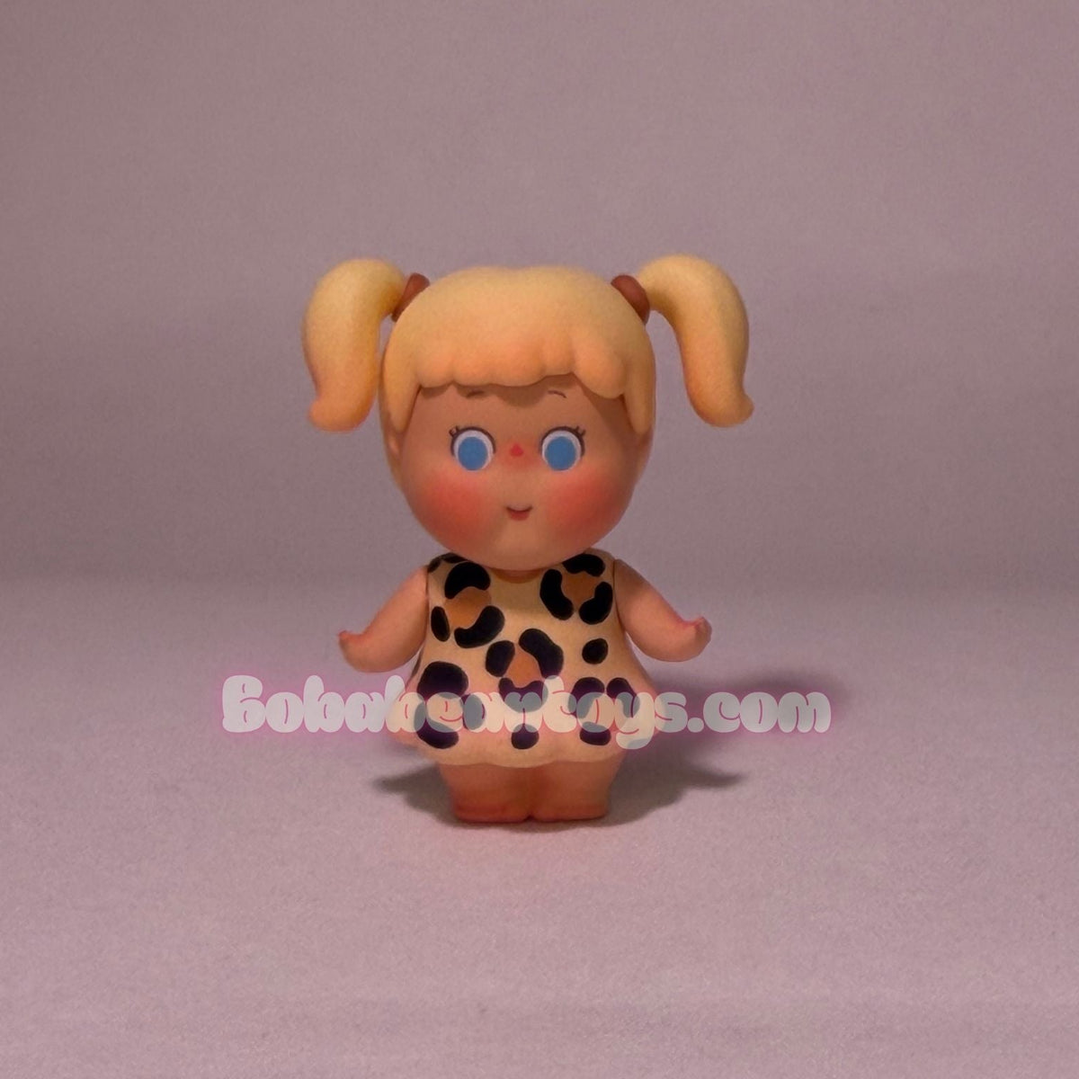A small vinyl doll with blonde pigtails, blue eyes, and a leopard-print dress, standing against a plain light gray background.