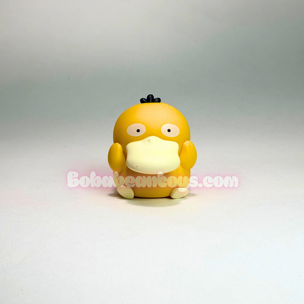 A small yellow duck-like toy figure with a cream-colored beak and mouth, standing against a plain light gray studio background. A pink watermark reading BobaBeastous.
