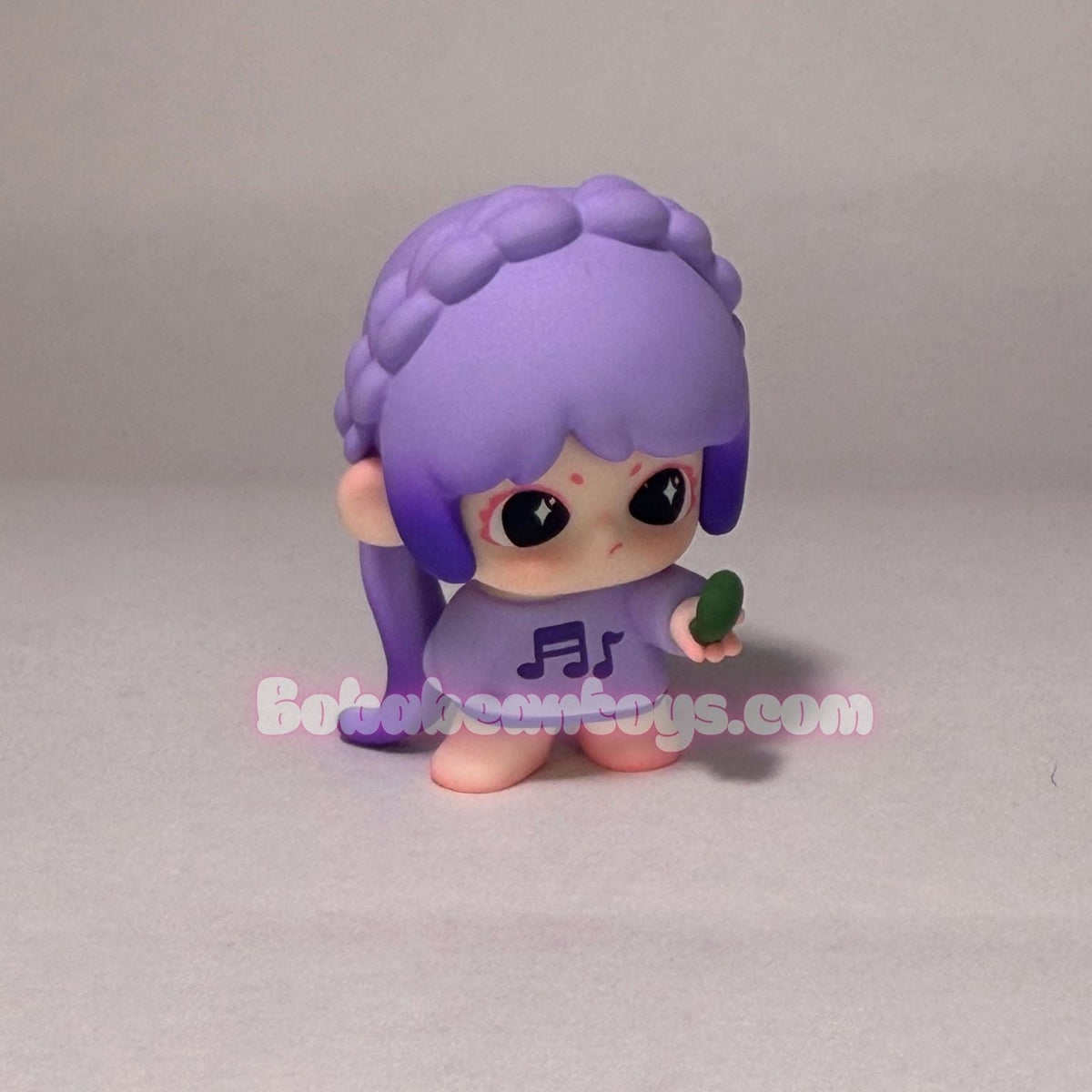 A small purple cartoon figurine with long braided hair, large black eyes, and a purple outfit, standing on a plain light gray background. A faint 'Botobin Toys.