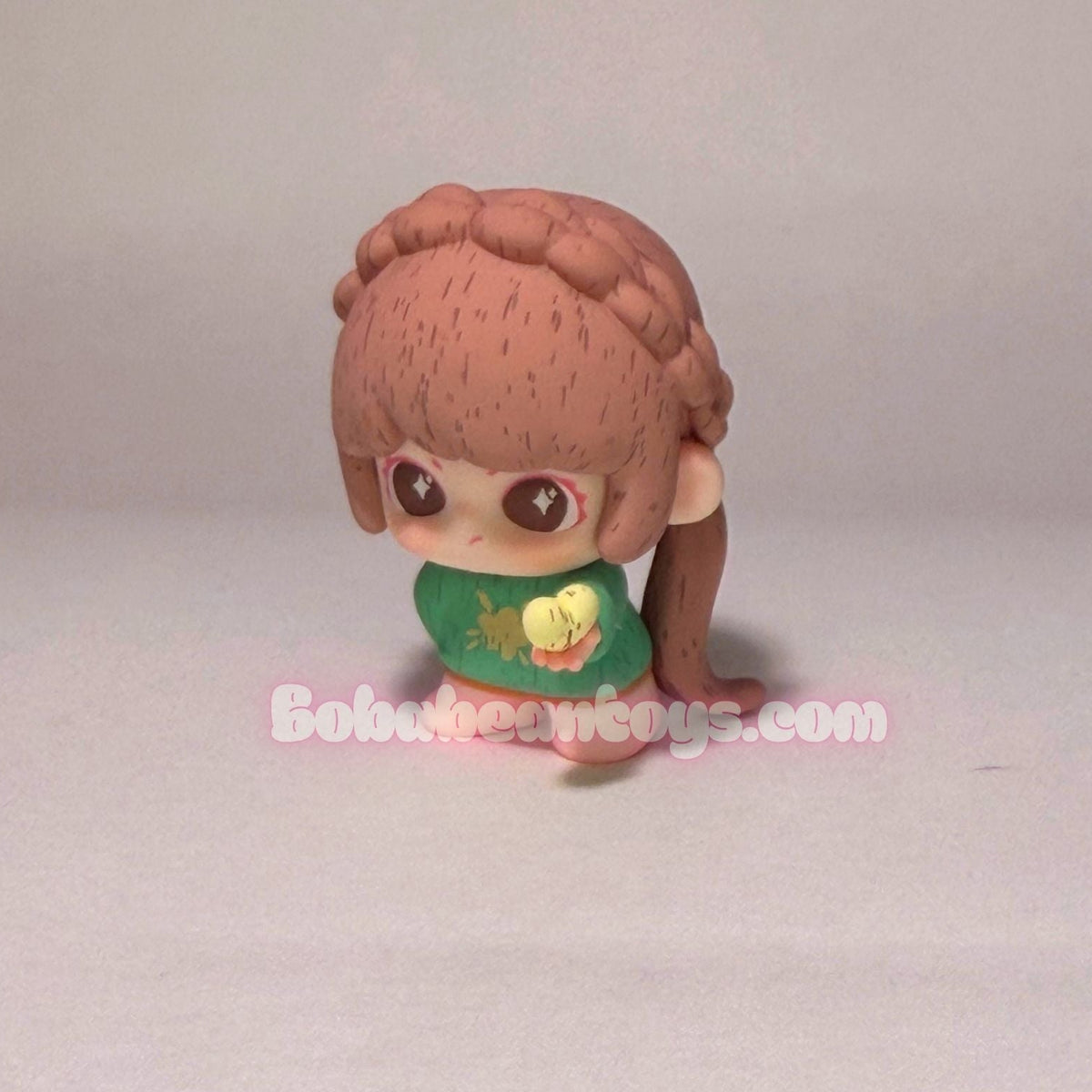 A small stylized doll figure with pinkish-brown hair, large eyes, and a green top with a yellow graphic, standing against a plain light background. A faint 'BobbeanToys.