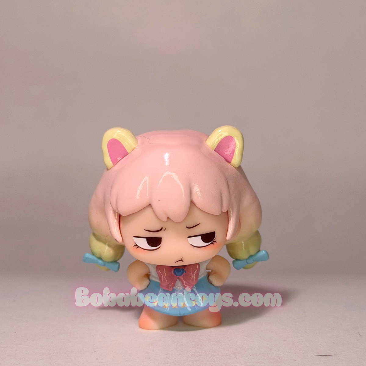 A small anime-style collectible figure with pink hair, yellow cat-ear-like tops, and a blue outfit, standing against a plain light gray background. A watermark reading BobaBeanToys.