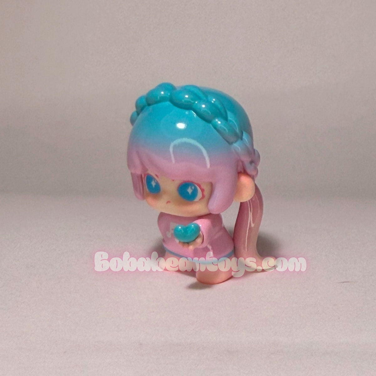 A small plastic doll figurine with pastel pink and blue hair, wearing a pink outfit and holding a blue heart, photographed against a plain light gray background.