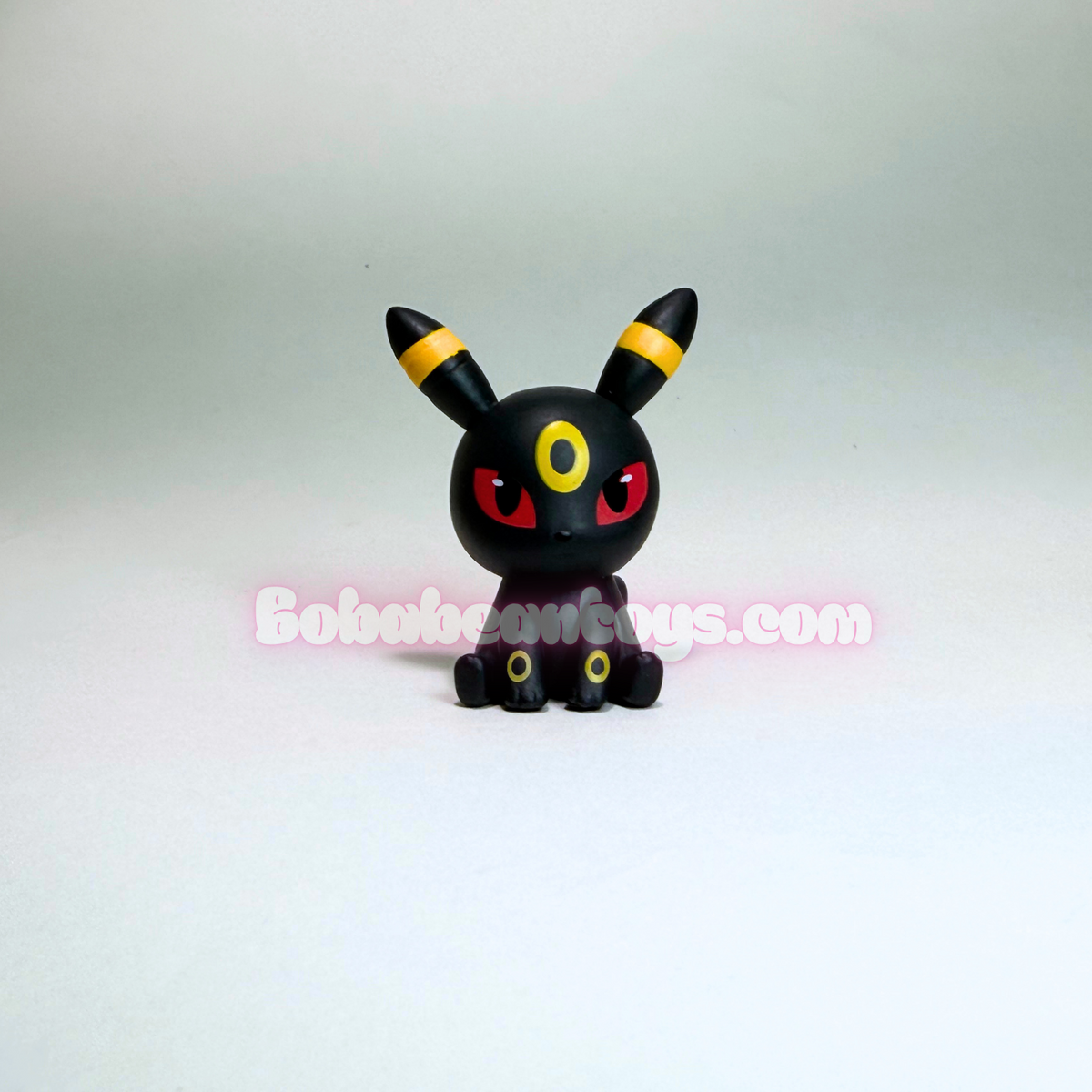 A small black Umbreon figurine with yellow-striped ears, red eyes, and yellow circular accents on its feet, centered against a plain light gray background. A pink watermark reading 'Bobooboy Toys.