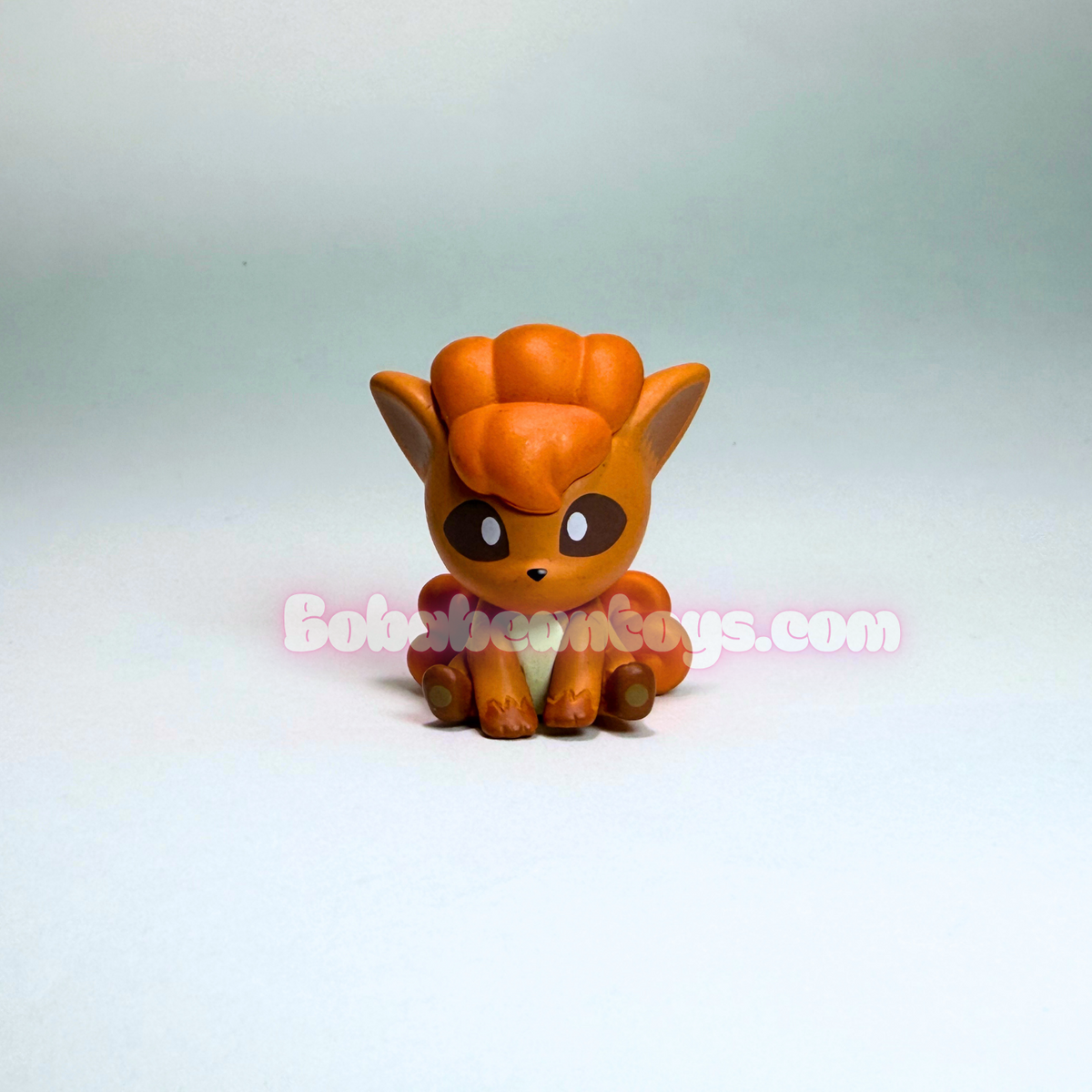 A small orange Pokémon figurine of a fox-like creature with large eyes, sitting on a light gray gradient background. A semi-transparent watermark reading 'BokokeToys.