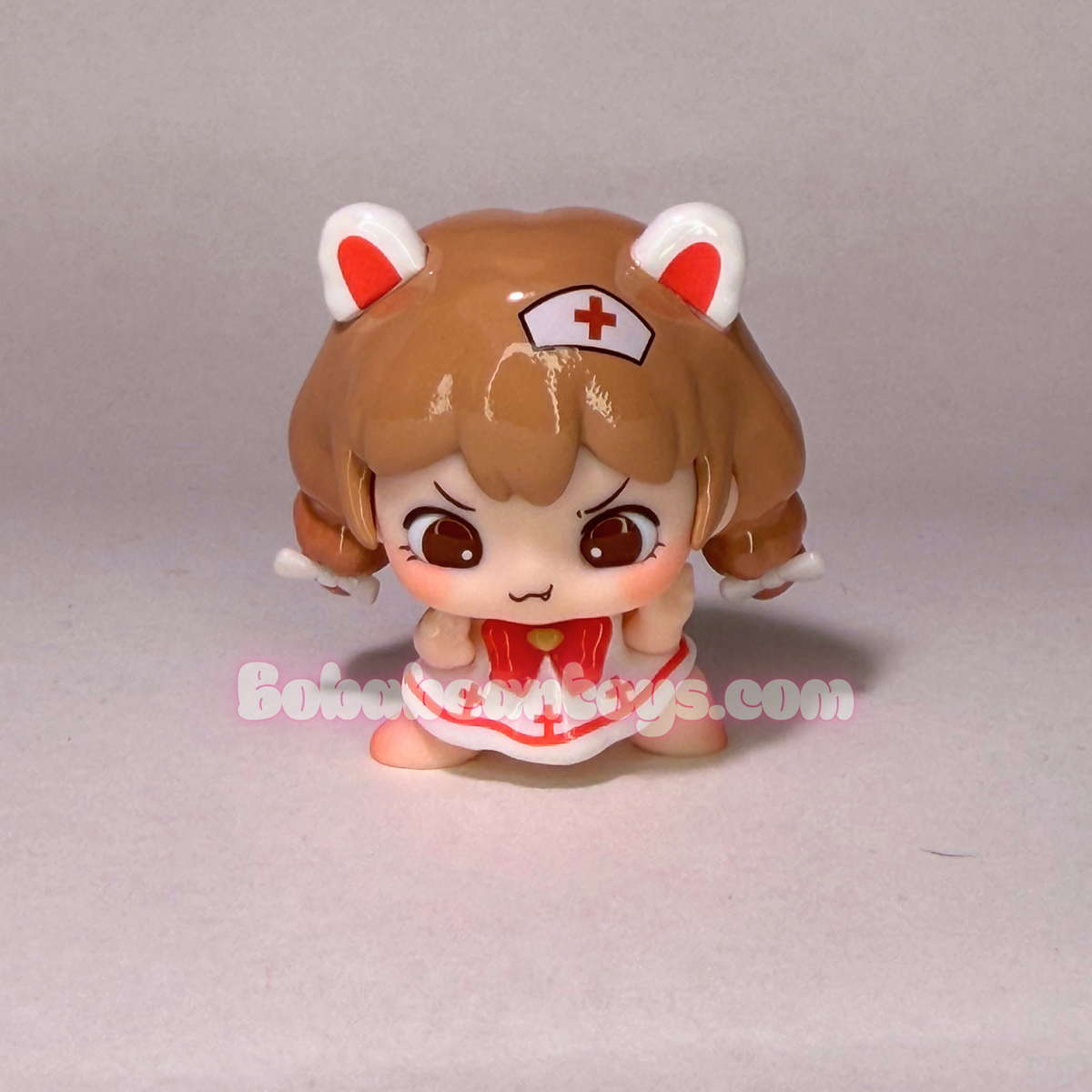 A small anime-style figurine of a brown-haired girl with cat-ear headphones and a nurse hat, wearing a red and white outfit, photographed against a plain light gray background.